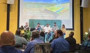 Panelists seated on stage at the 2026 WHIN Ag Summit at the Beck Agricultural Center, discussing Intelinair in front of a projected slide featuring the Intelinair logo, aerial field imagery, and the quote 'Farmers won't touch a field decision without checking Intelinair.'
